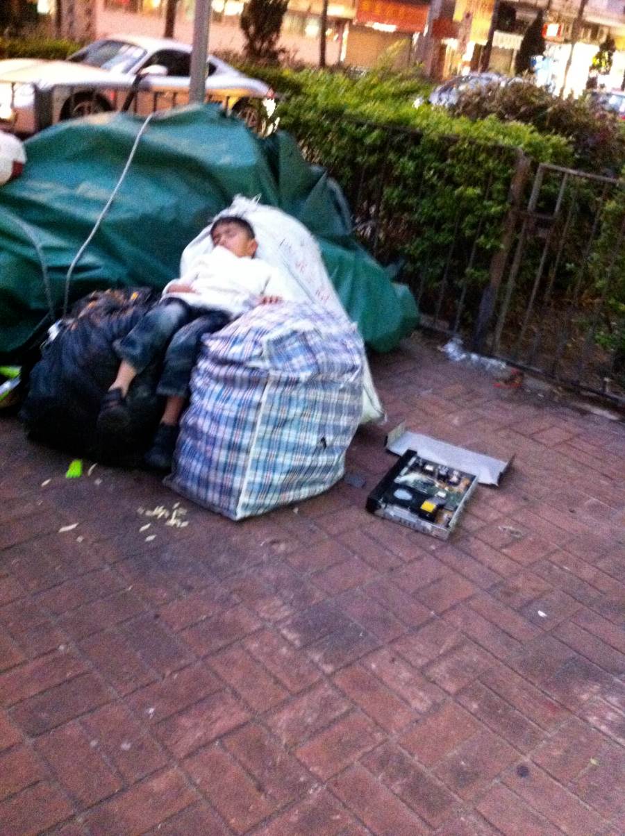 He doesn't actually live here but this traffic island is one of a type of recycling plant that crop up everywhere. His dad/uncles collect rubbish, store it here and earn money from the valuable parts such as wire and metal. Sham Shui Po is recognised as a destination for migrant workers historically and in the present. This boy's family speak Cantonese so they may have grown up here.